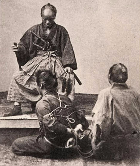 Three men in hojojutsu training, in which one man is tying th erestraints, another is being bound and a third is explaining / teaching the practice.
