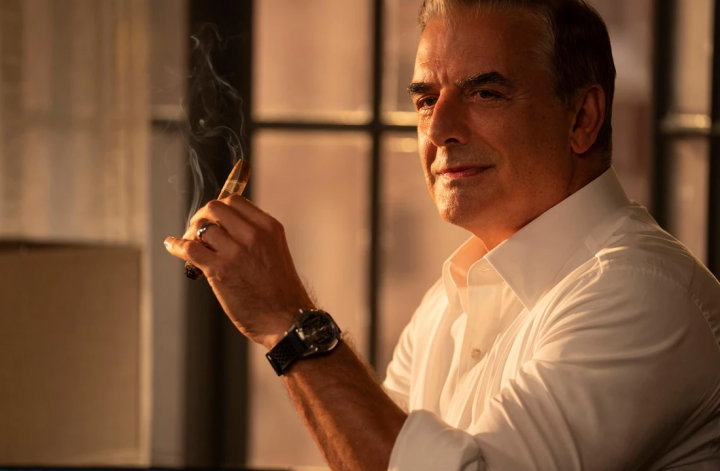 Image of Chris Noth from And Just Like That, holding a cigar and looking out of a window at the sunset